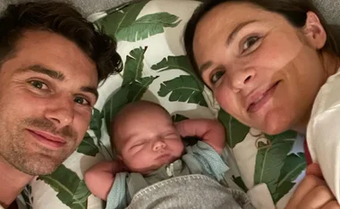 Family selfie with a sleeping baby between smiling parents on a leafy-patterned blanket.