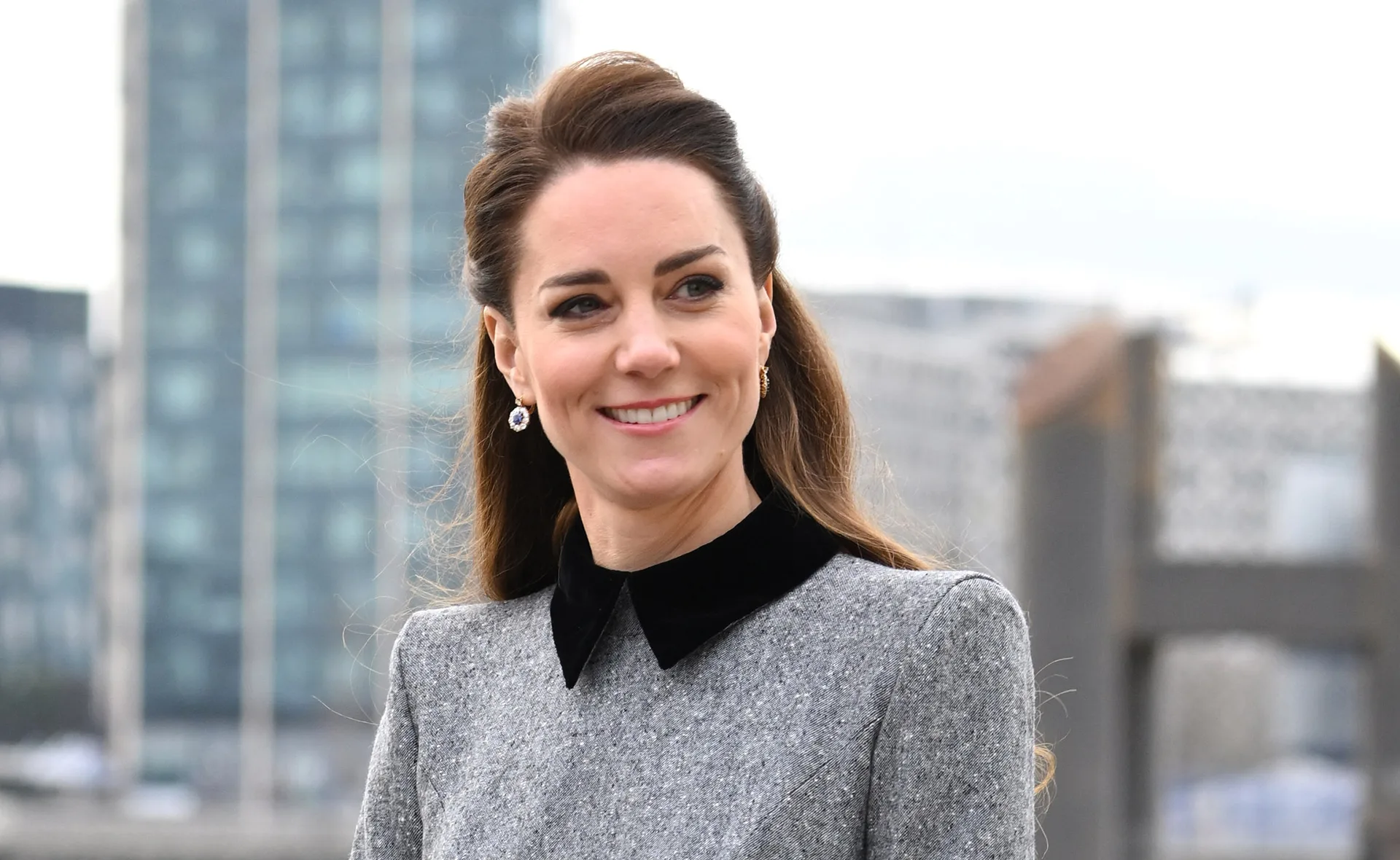 A woman in a grey coat dress with a black collar and earrings, smiling in an outdoor urban setting.
