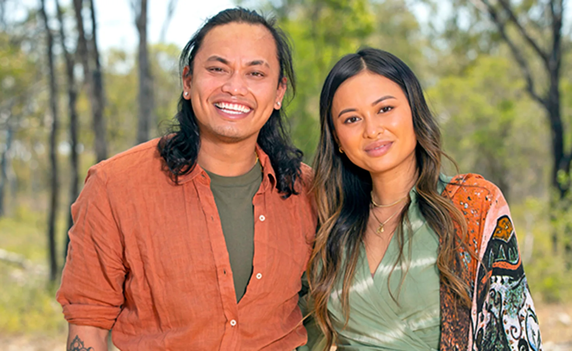 Two people smiling outdoors, with trees in the background; one in an orange shirt, the other in a green top with a shawl.