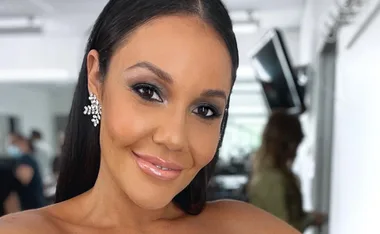 Close-up of a smiling woman with long dark hair and sparkly earrings in a bright indoor setting.