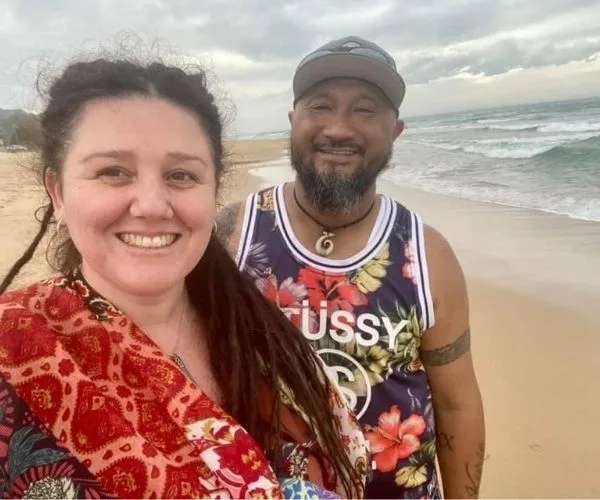 A smiling couple takes a selfie on a beach, with waves in the background and cloudy skies above.
