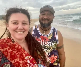 A smiling couple takes a selfie on a beach, with waves in the background and cloudy skies above.