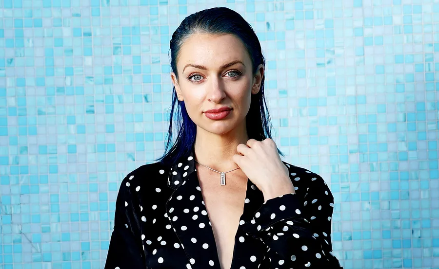 Portrait of a person with slicked-back hair and polka dot attire against a blue tiled background.