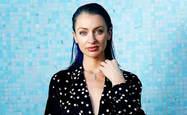 Portrait of a person with slicked-back hair and polka dot attire against a blue tiled background.