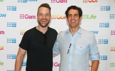 Two men smiling in front of a backdrop with colorful TV network logos.