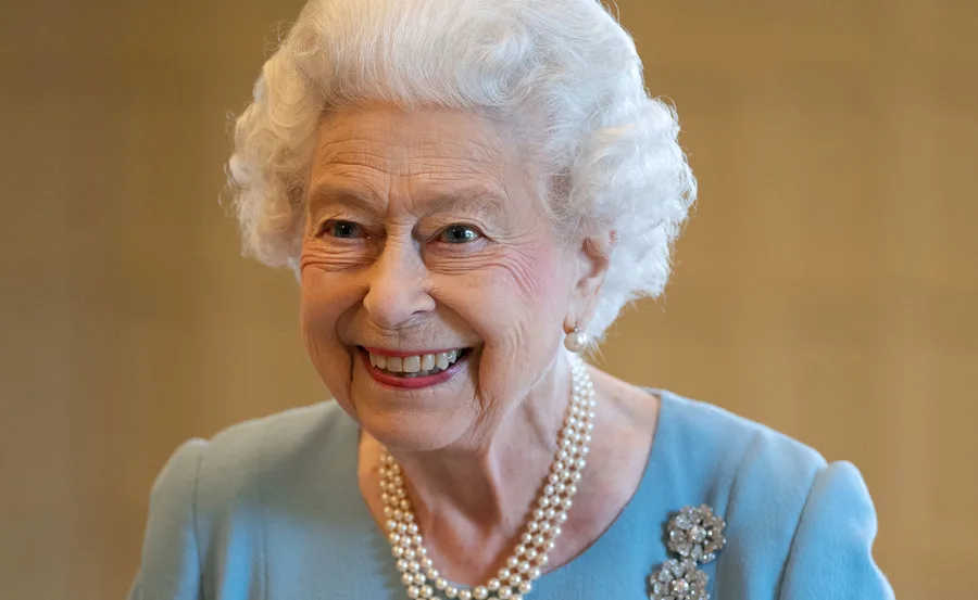 A smiling elderly woman with white hair, wearing pearls and a light blue outfit.