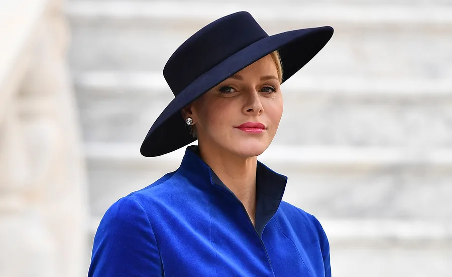 Woman in a blue coat and black wide-brimmed hat, standing with a neutral expression.