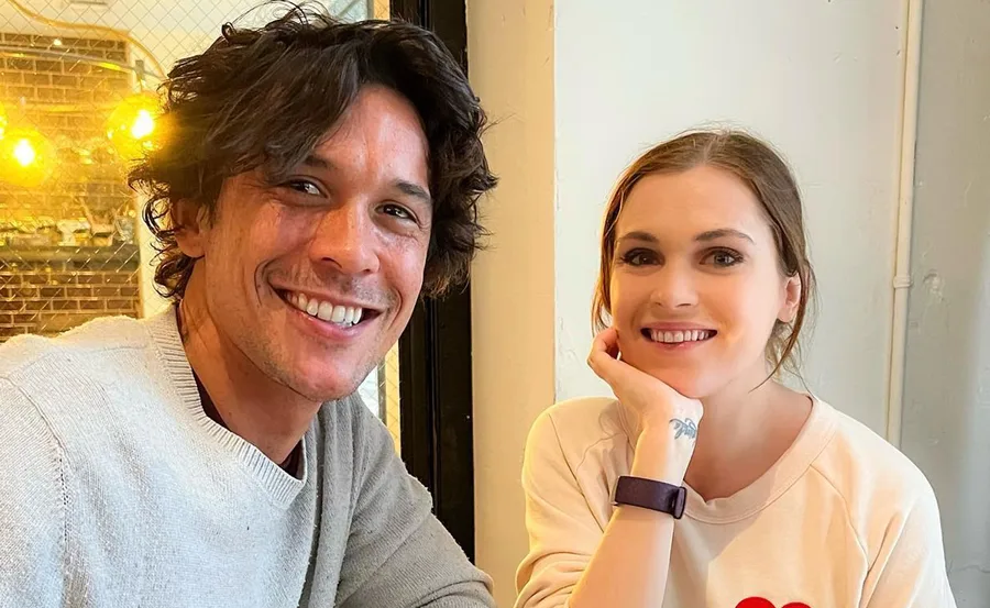 Two people smiling at a camera in a warmly lit indoor setting, sitting close together.