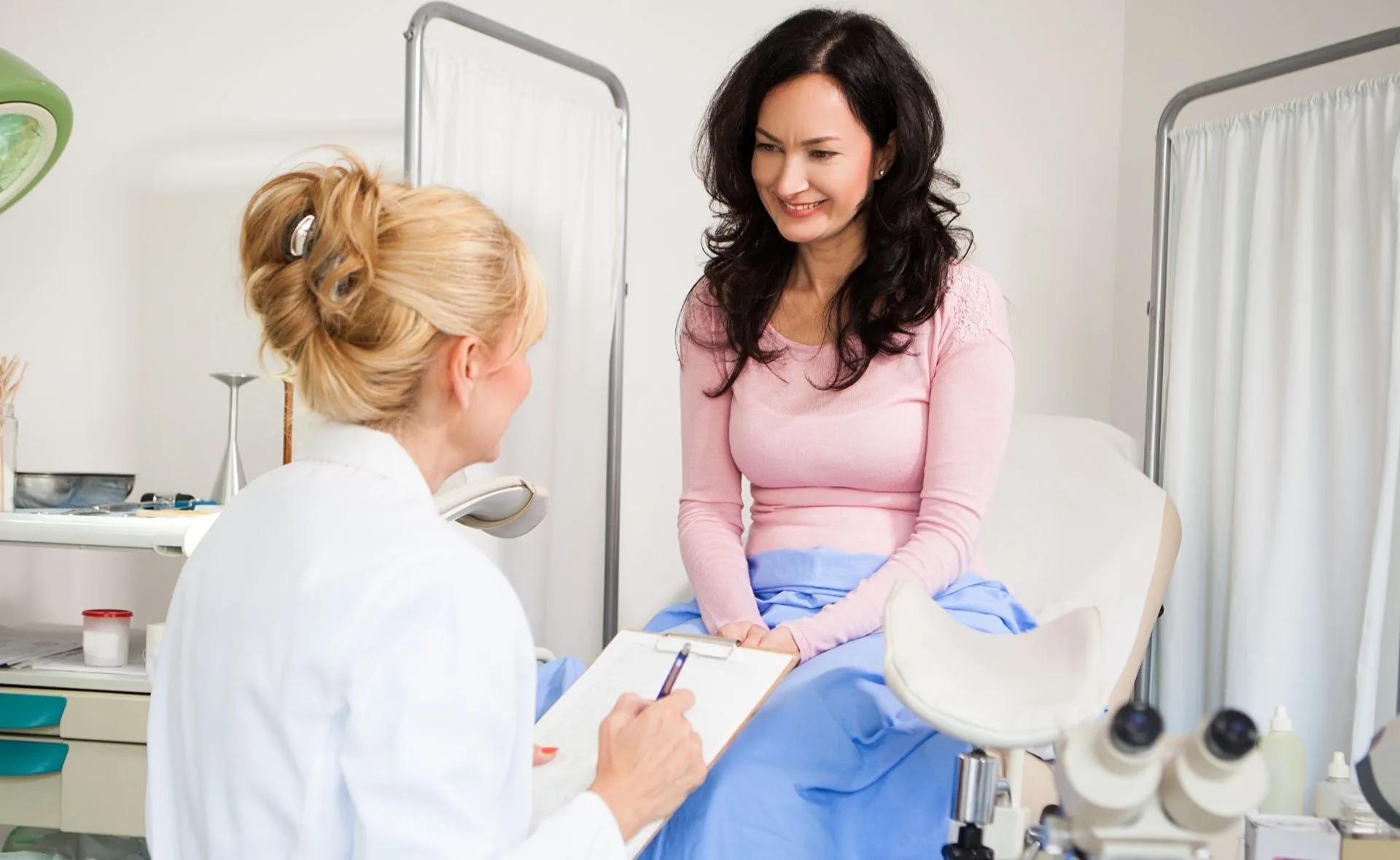 A woman sits on a medical examination table, smiling at a healthcare professional holding a clipboard.
