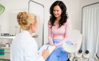 A woman sits on a medical examination table, smiling at a healthcare professional holding a clipboard.
