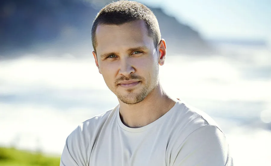A person with short hair and a light shirt stands outdoors with a blurred natural background.