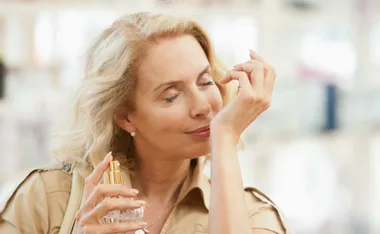 Elderly woman enjoying perfume scent on her wrist, holding a gold-capped bottle, eyes closed, smiling softly.