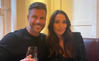 A smiling man and woman sit together on a couch, the man holding a wine glass.