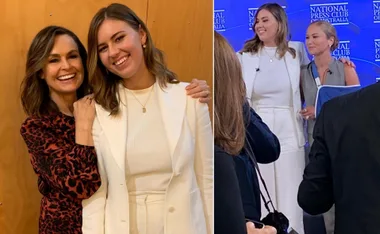 Two women smiling for a photo at the National Press Club, one in a patterned dress, another in a white outfit.