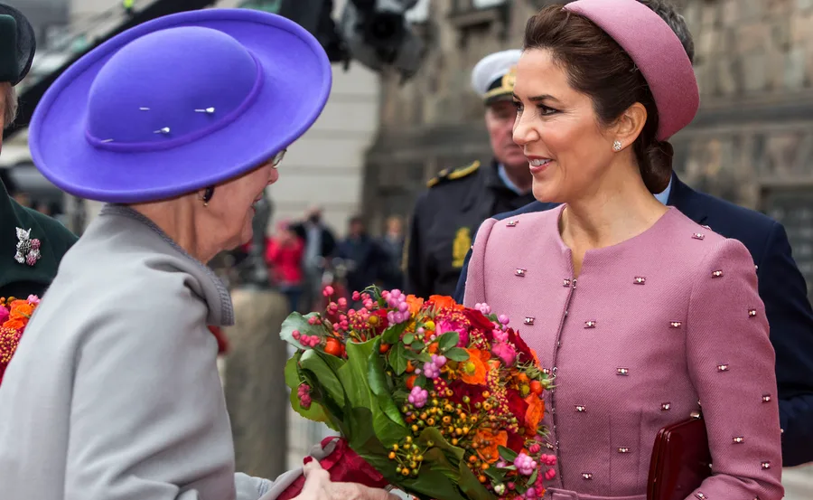 Two women exchanging flowers, one in a pink outfit, the other in a purple hat, with a background of onlookers and officers.