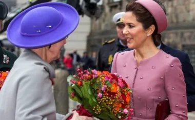 Two women exchanging flowers, one in a pink outfit, the other in a purple hat, with a background of onlookers and officers.