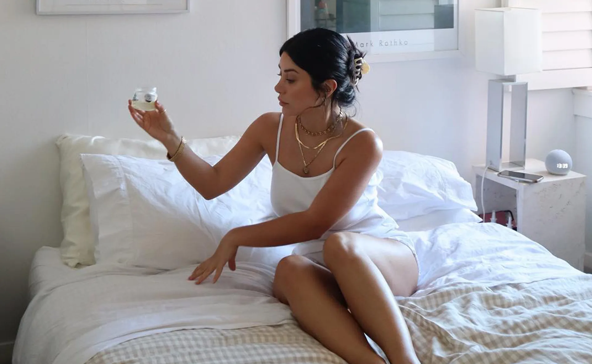 Woman in white outfit sitting on a bed holding a jar, beside a nightstand with a lamp and round clock.