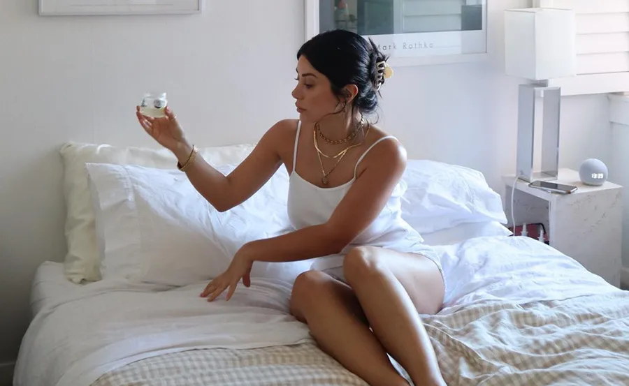Woman in white outfit sitting on a bed holding a jar, beside a nightstand with a lamp and round clock.