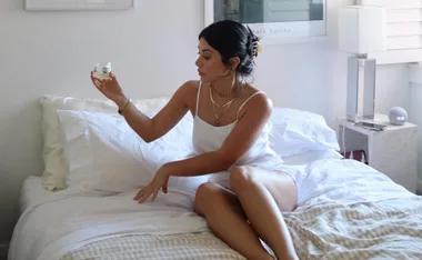 Woman in white outfit sitting on a bed holding a jar, beside a nightstand with a lamp and round clock.