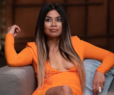 A person in an orange dress sits on a couch with a thoughtful expression.