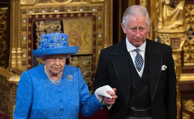 Elderly woman in blue outfit and hat holding hands with a man in a black suit, both in a richly decorated room.