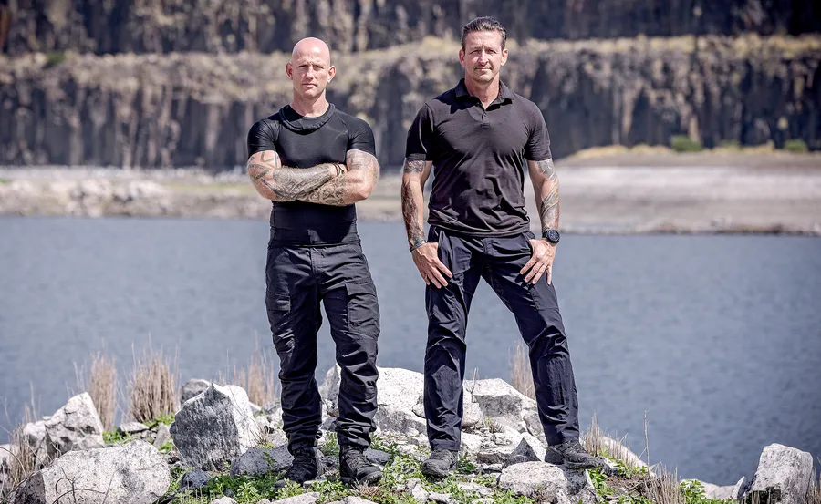 Two men in tactical clothing stand on rocky terrain by a lake, with cliffs in the background.