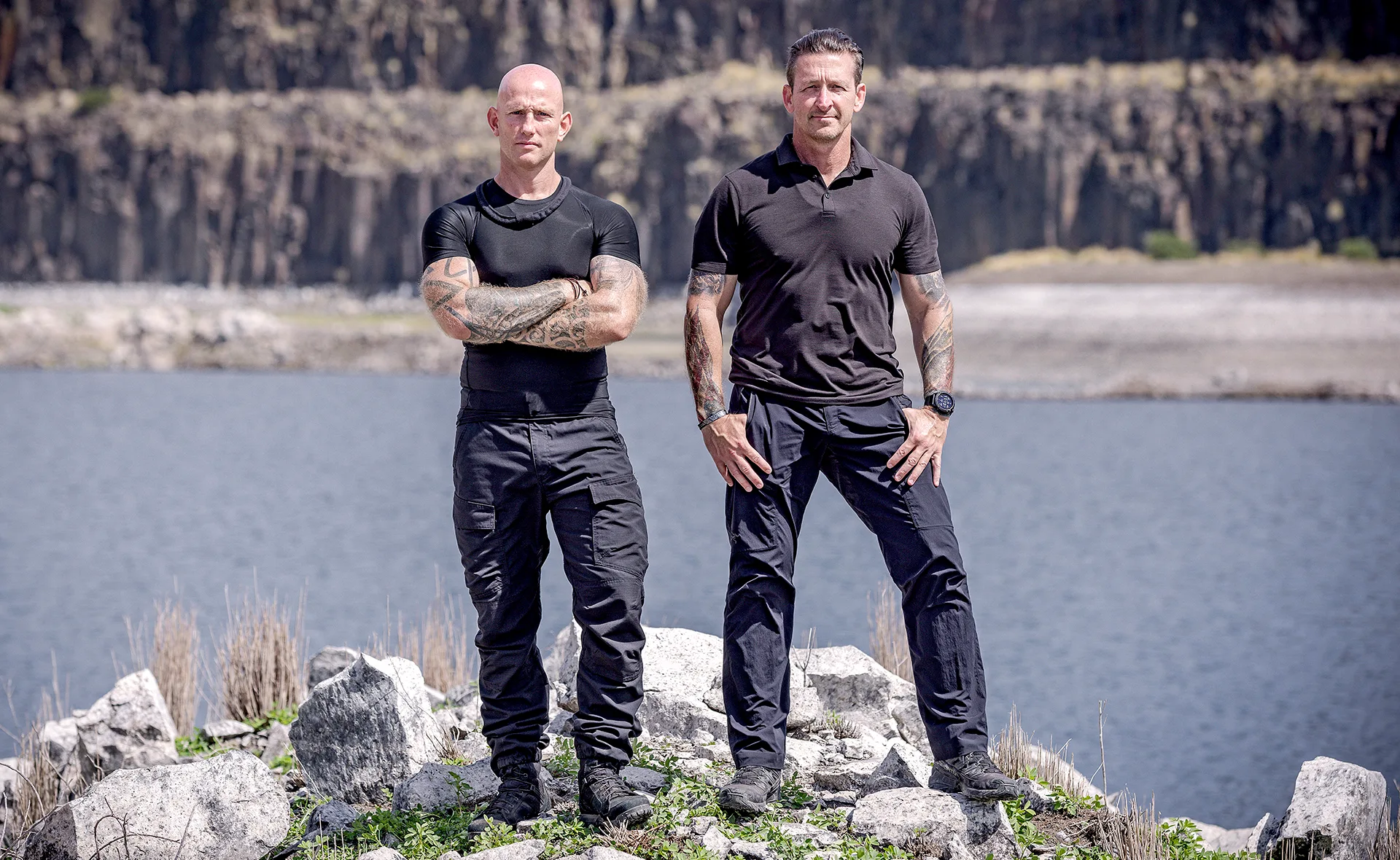 Two men in tactical clothing stand on rocky terrain by a lake, with cliffs in the background.