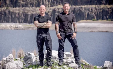 Two men in tactical clothing stand on rocky terrain by a lake, with cliffs in the background.