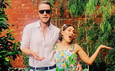 Man in sunglasses with woman in colorful dress posing playfully in front of brick wall and greenery.
