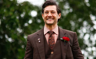 Smiling man in brown suit with red rose boutonnière and patterned tie, set against a blurred green background.