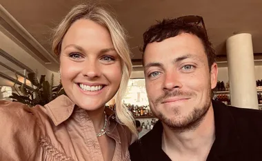 Two people smiling at the camera in a cozy indoor setting with plants in the background.