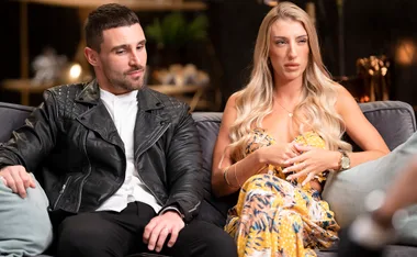Man in black leather jacket and woman in patterned dress sitting on a couch, engaged in conversation.