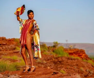 Person in colorful robe and scarf poses confidently with fan, standing on rocky terrain in sunny landscape.
