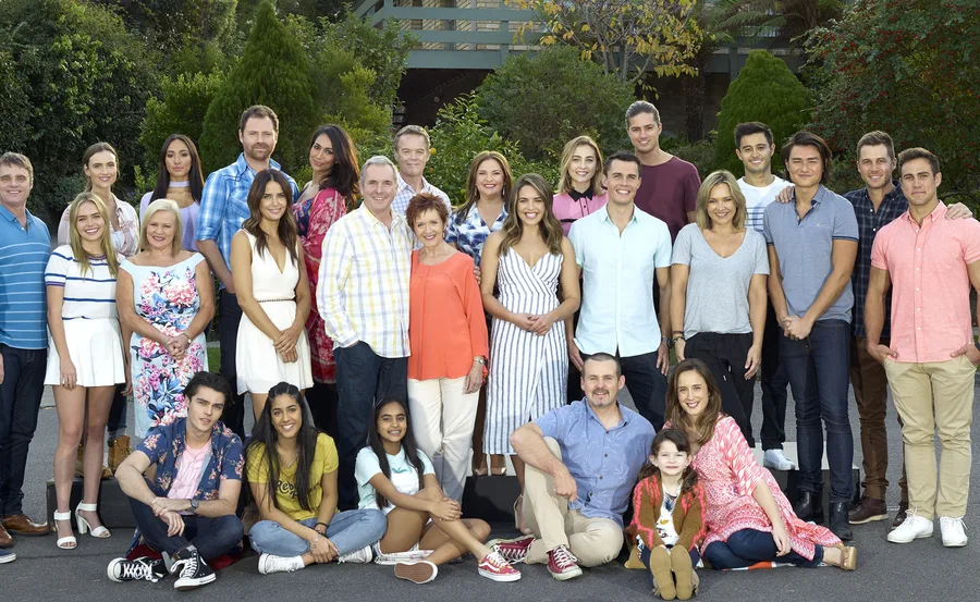 A large group of people, smiling and standing outdoors, with greenery in the background.