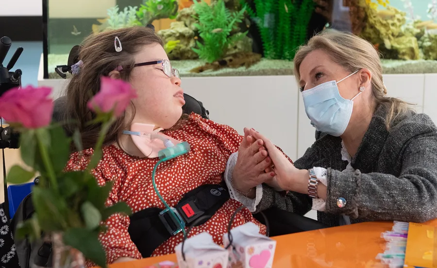 A masked woman holds hands with a girl in a wheelchair at a table with pink roses.