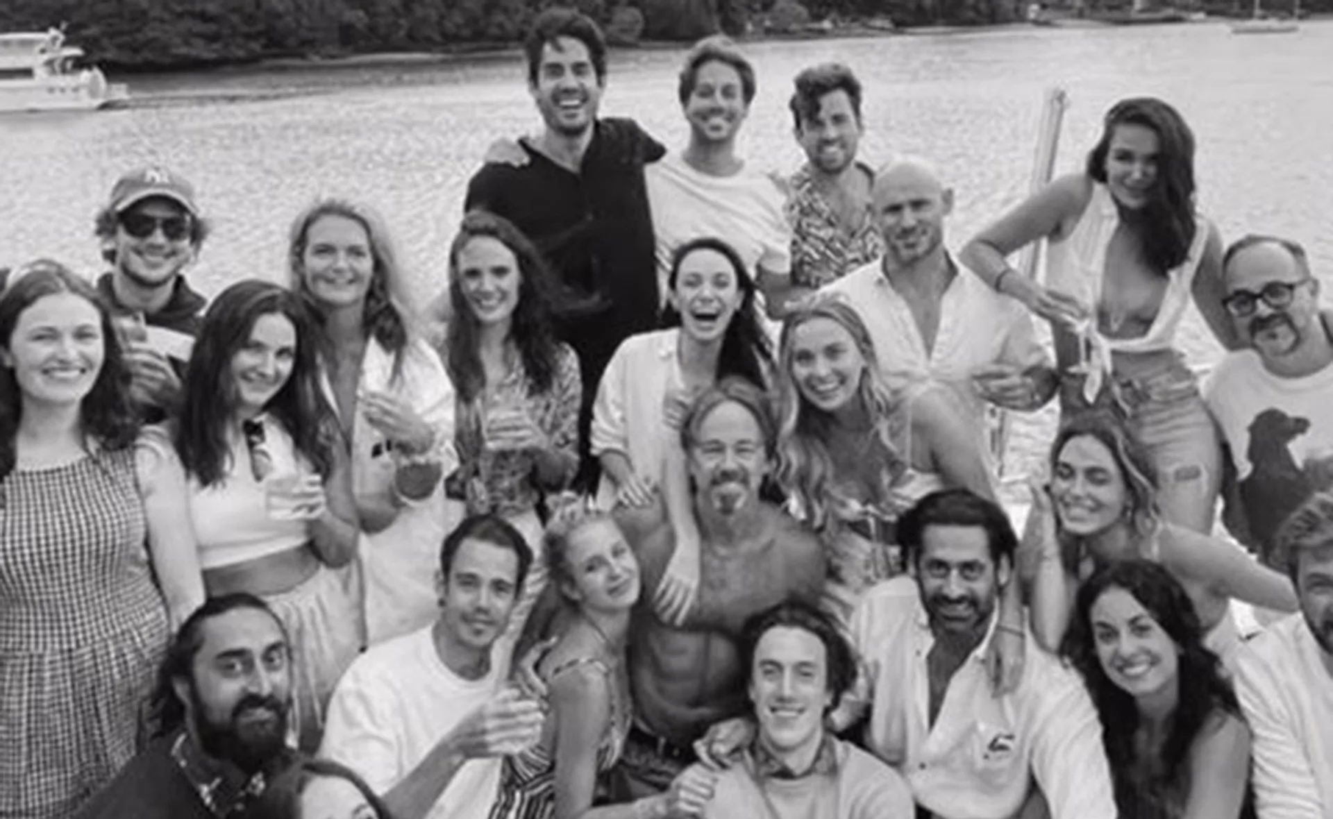 Group of friends smiling and posing on a boat, with a scenic river view in the background, in black and white.