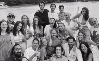 Group of friends smiling and posing on a boat, with a scenic river view in the background, in black and white.