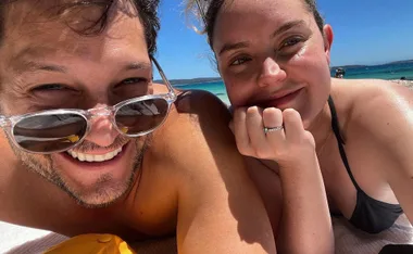 A couple smiling close together, lying on a beach with clear blue skies and ocean in the background.