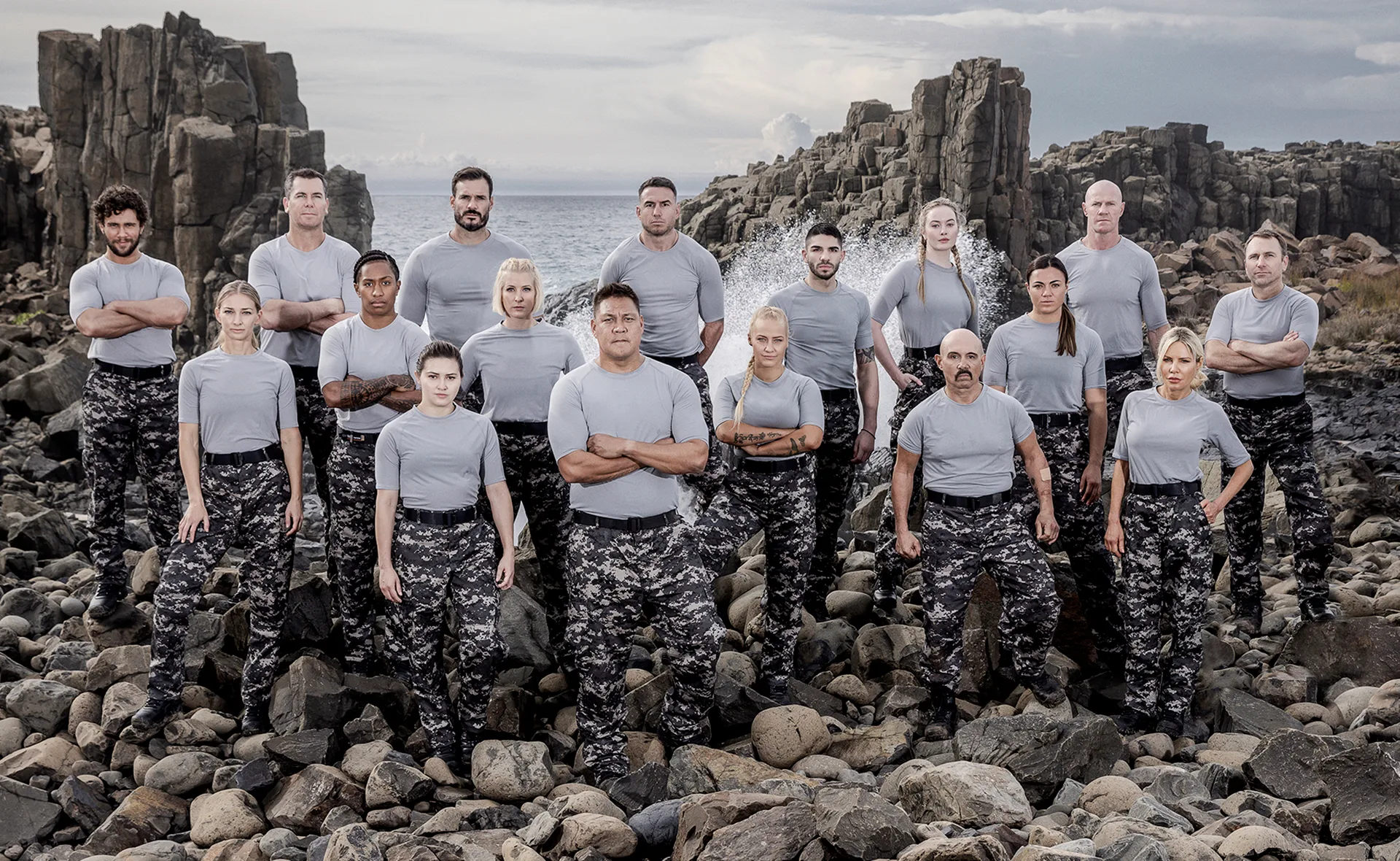 Contestants of SAS Australia stand on rocky terrain in grey shirts and camo pants, filmed at a coastal location.