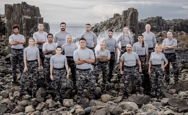 Contestants of SAS Australia stand on rocky terrain in grey shirts and camo pants, filmed at a coastal location.