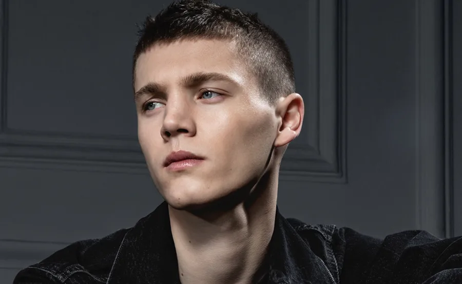 Young man with short hair in a contemplative pose, wearing a dark jacket, against a muted background.