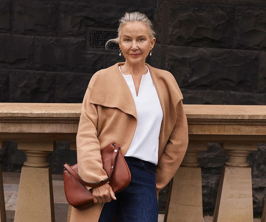 Senior woman in camel coat and white blouse with brown leather bag, standing by stone railing.