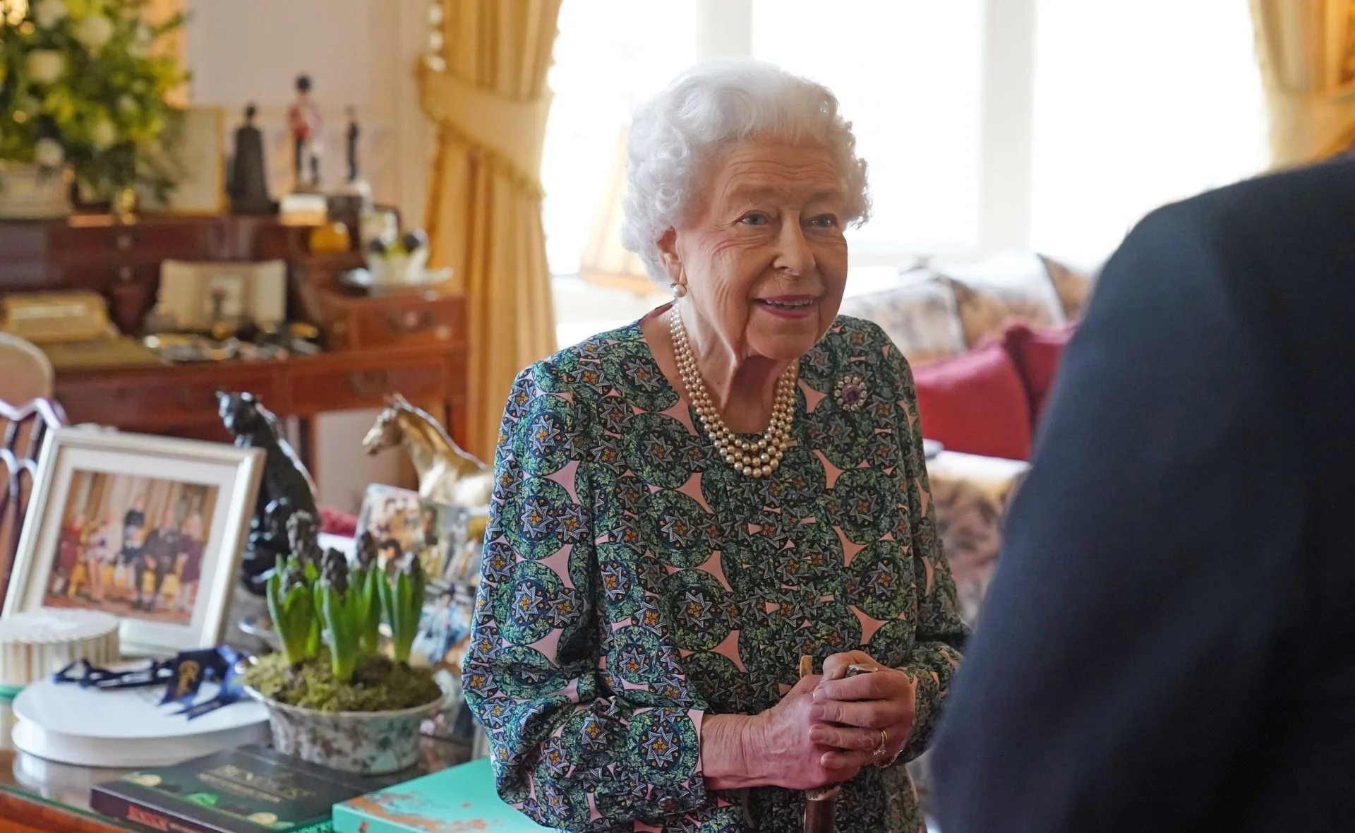 Elderly woman in a patterned green dress holding a cane, standing in a room with photos and decor on a table.