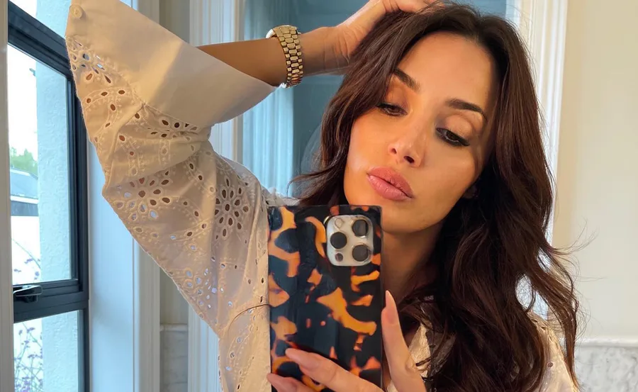 Woman with long brown hair taking a selfie in a mirror, wearing a white eyelet blouse.