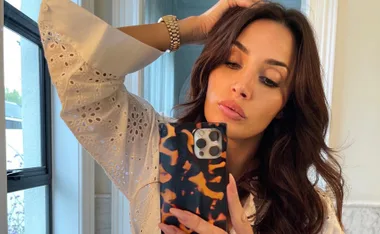 Woman with long brown hair taking a selfie in a mirror, wearing a white eyelet blouse.