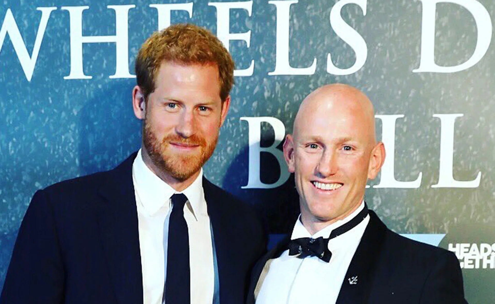 Two men in suits smiling at an event, with blue background and white text partially visible.