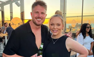 A couple smiles in a sunset-lit venue, with guests and ocean view in the background.