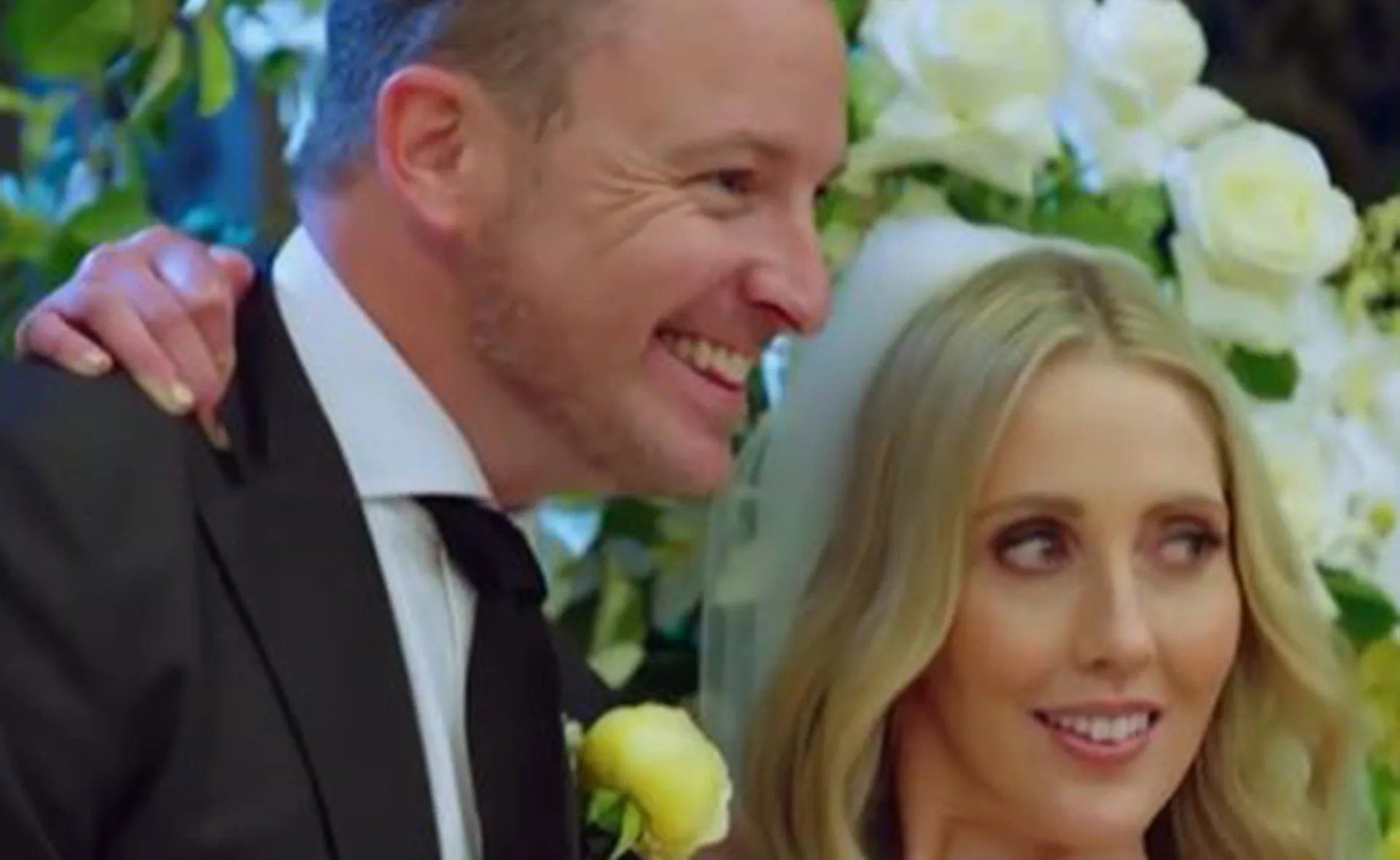 A smiling couple at a wedding, with white roses in the background.