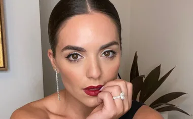 A woman with slicked-back hair, red lipstick, long earrings, and a large ring, posing thoughtfully with her hand on her chin.