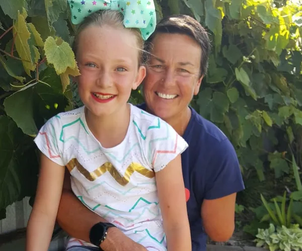 A smiling woman and young girl with a bow, sitting together outdoors among green leaves.